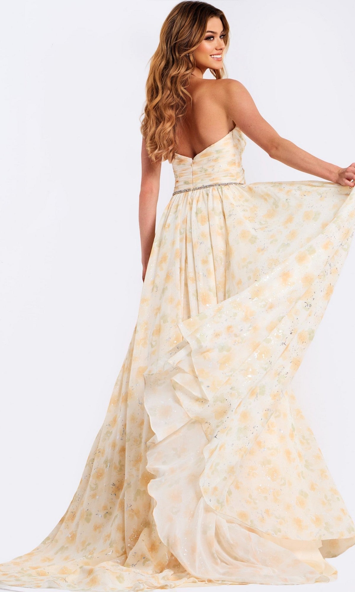 A woman in the Jovani 48139 long prom dress holds out the skirt, highlighting its strapless chiffon design and flowing fabric with a subtle pale yellow floral-print pattern.